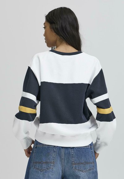Pulover colorat în blocuri de navy, alb și galben, cu textură ribbed și mâneci largi. Dispune de un guler rotund și tiv elastic.