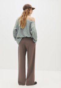 Femme portant un pull gris à épaules dénudées, un pantalon large marron, des chaussures noires et une casquette marron, se tenant devant un fond blanc uni.