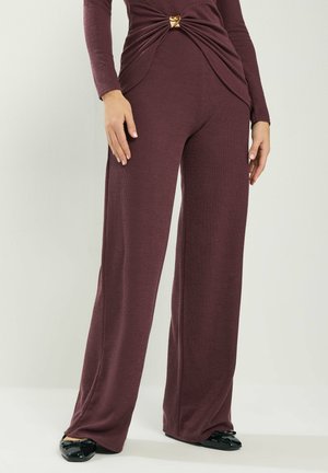 Kvinde iført bordeaux strikkede bukser med vide ben og matchende top med snoet detalje, fastgjort med en guldlås, kombineret med sorte lakflade sko.