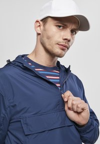 Tengerész kék zipzáras dzseki, elülső zsebbel, könnyű anyagból, állítható kapucnival, csíkos kék és piros póló felett viselve, amit egy fehér sapka egészít ki.