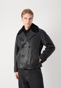 Musta nahkatakki, jossa on kaksirivinen muotoilu, turkista tehty kaulus ja sivutasku vetoketjulla. Silkkinen rakenne erottuvilla napilla ja istuvalla leikkauksella.