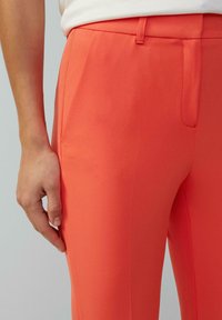 Korallefarbene Hose mit glatter Textur, die eine taillierte Passform, seitliche Taschen und ein flaches Vorderdesign aufweist.