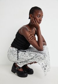 Haut noir texturé avec des bretelles en chaîne, jupe argentée à sequins brillants, et chaussures noires à talons épais avec nœuds et accents décoratifs.
