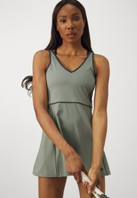 Vestido atlético verde com decote em V, corpete ajustado, saia ampla, tecido suave e detalhes em contraste, usado por uma pessoa segurando uma raquete de ténis.