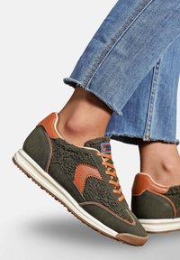 Des baskets vertes avec des accents orange, un dessus texturé, une semelle blanche et des lacets contrastants. Portées avec un jean bleu effiloché.