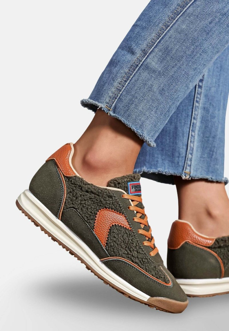 Des baskets vertes avec des accents orange, un dessus texturé, une semelle blanche et des lacets contrastants. Portées avec un jean bleu effiloché.