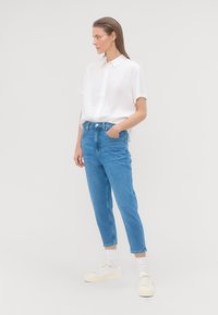 Žena stojí s jednou rukou vo vrecku, oblečená v bielej tričko s krátkym rukávom, modrých džínsoch, bielych ponožkách a bielych teniskách, na jednofarebnom pozadí.