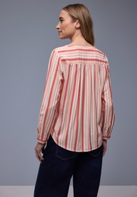 Femme de profil portant une blouse à manches longues avec des rayures verticales rouges, pêche et blanches, et un jean bleu foncé sur un fond gris.