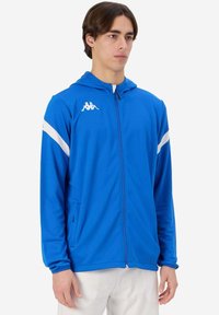 Felpa blu con zip realizzata in tessuto liscio, caratterizzata da una striscia bianca sulle spalle e un logo sul lato sinistro del petto. Due tasche laterali.