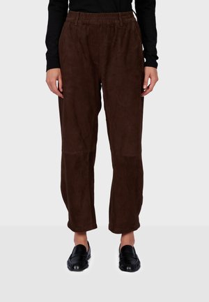 Personne portant un pantalon en daim brun foncé avec une taille élastique et des mocassins noirs, debout sur un fond clair uni.