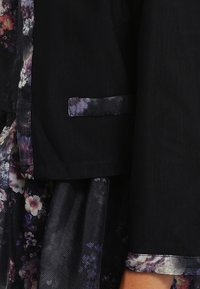 Svart jacka med ett blommönstrat foder, med vävt mönster och en framficka. Kragen och ärmsluten har en glänsande yta.