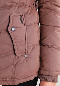 Brown puffer jacket with a textured finish, featuring a snap pocket, visible stitching, and ribbed cuffs for warmth.