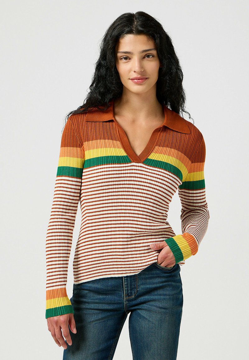 Donna con lunghi capelli ricci che indossa un maglione a coste con strisce marroni, arancioni, gialle e verdi, abbinato a jeans blu, con una mano in tasca.