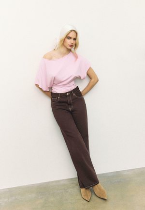 Žena naslonjena na zid nosi blijedosmeđu bluzu bez ramena, tamnosmeđe široke hlače, bež špičaste čizme i bijeli povez za glavu.