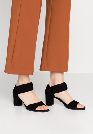 Füße, die schwarze Sandalen mit Blockabsatz und elastischen Knöchelriemen tragen, kombiniert mit braunen Hosen mit weitem Bein vor hellem Hintergrund.