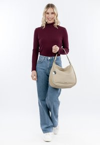 Beige leather handbag with a curved shape, silver hardware, and a short strap. Model wears a maroon ribbed top and light blue wide-leg jeans.