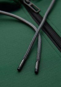 Giacca verde con zip, caratterizzata da una cerniera nera e passanti a cordino texturizzati con motivo bianco e nero. Gli accessori includono punte nere con logo.