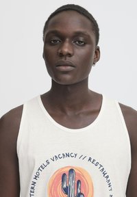 Balts tanktops ar kaktusa grafisko dizainu un apļveida akcentiem oranžā krāsā, kā arī zilā tekstā. Modelis ar īsiem matiem un auskaru.