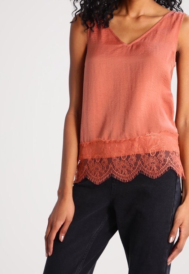 Brzoskwiniowy top na ramiączkach w serek z teksturowanym wykończeniem i koronkowym wykończeniem na dolnej krawędzi; zestawiony z ciemnymi spodniami, prezentując swobodny styl.