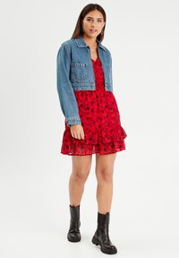 Giacca di jeans cropped sopra un vestito floreale rosso con orlo arricciato. Modella che indossa stivali neri alla caviglia, esibendo tessuti lisci e colori vivaci.
