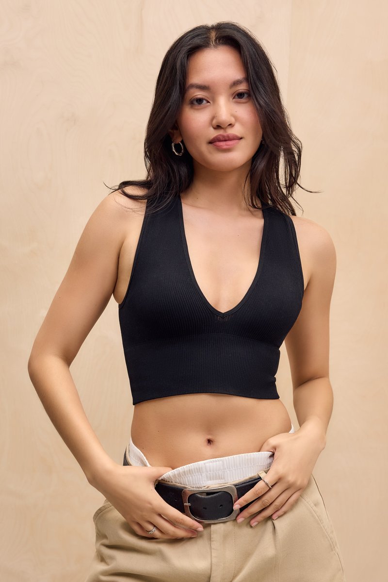 Junge Frau mit langen dunklen Haaren, die ein schwarzes, ärmelloses Crop-Top und beige Hosen trägt und einen schwarzen Gürtel anpasst, vor einem neutralen Holz-Hintergrund.