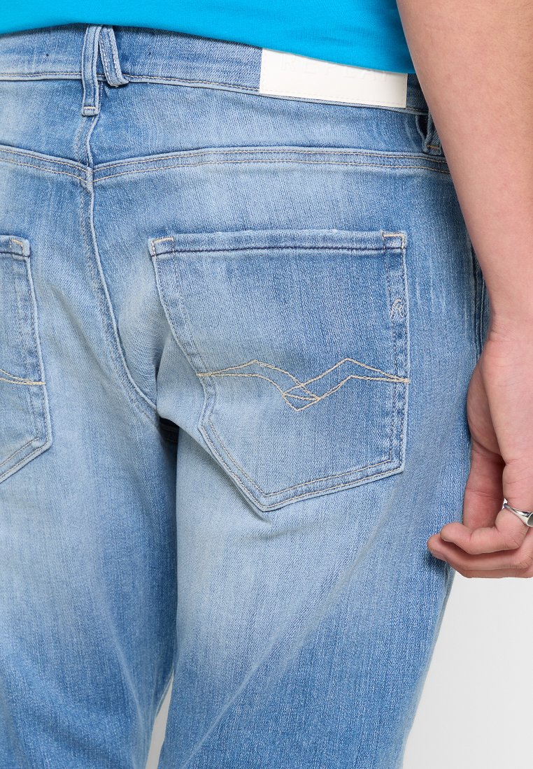 Hellblaue Denim-Jeans mit einem ausgewaschenen Finish, ausgestattet mit zwei Gesäßtaschen und tonalem Nähmuster im Zickzack. Weißes Taillenband-Detail sichtbar.