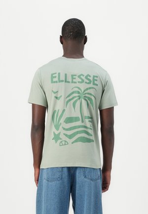 Homme portant un t-shirt Ellesse vert clair avec des motifs de palmier et de plage dans le dos, assorti à un jean bleu, vu de dos.