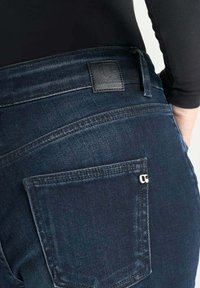 Jean en denim bleu foncé avec une pièce sur la taille arrière, coutures visibles et un petit détail de poche zippée à l'arrière.