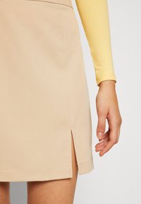 Falda corta beige con una abertura lateral y una textura suave. La prenda se combina con una blusa amarilla de manga larga con relieve.