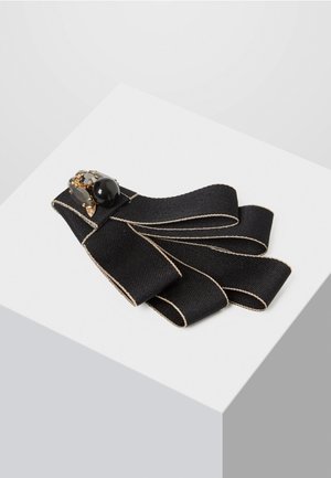 Černá látková brož s béžovými okraji, složená do smyček, se sponou zdobenou černými a jantarovými kameny.