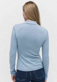 Ljusblå långärmad polo t-shirt, mjukt stickat material, med krage och ribbade ärmslut. Bärs med mörka denimjeans.