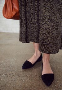 Flats pointues en daim noir à coupe basse, associées à une robe noire à motif de pois jaunes, et un sac à main en cuir marron.