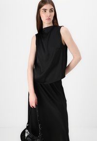 Juoda beapykaklė suknelė su aukštu kaklu, laisva siluetu ir glotnia, blizgia medžiaga. Modelis laiko juodą rankinę su blizgiais akcentais.