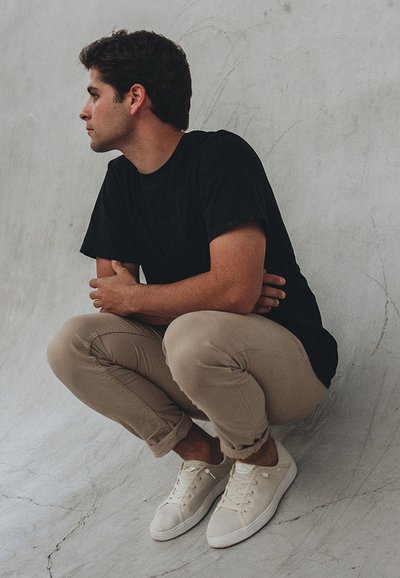 Zwarte t-shirt, beige broek en witte sneakers. Onderwerp zit in een squat tegen een textuur achtergrond in grijs, met de armen gekruist op de knieën.