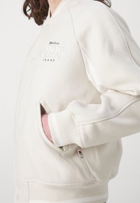 Biela bomber bunda so ribovanými manžetami a spodným lemom. Obsahuje vyšívané logo "Tommy Jeans" a bočné vrecká. Mäkká textúra.