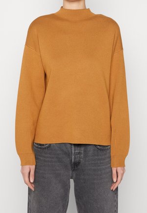 Osoba ubrana w musztardowożółty sweter z długim rękawem i wyblakłe czarne dżinsy, stojąca na tle jednolitego białego tła.