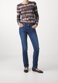 Donna che indossa jeans dritti blu, una maglietta a manica lunga nera e beige con motivo tie-dye e ballerine nere, con le mani nelle tasche.