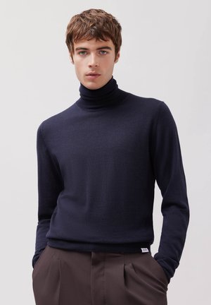 Ung mand med kort brunt hår iført en mørk marineblå rullekrave og brune bukser, stående med hænderne i lommerne mod en ensfarvet baggrund.