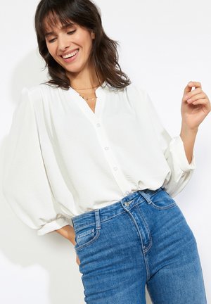 Kobieta uśmiecha się, ubrana w luźną białą koszulę na guziki z bufiastymi rękawami i niebieskie dżinsy z wysokim stanem, pozująca na białym tle.