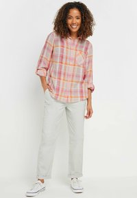 Camicia a scacchi nei colori rosa, arancione e crema, con bottoni sul davanti, maniche lunghe arrotolate fino ai gomiti, abbinata a pantaloni a sigaretta chiari e sneakers bianche.