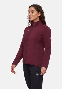 Burgundowa polarowa kurtka z wysokim kołnierzem, pełnym zamkiem błyskawicznym i kieszeniami po bokach. Zawiera logo na klatce piersiowej oraz naszywkę na lewym rękawie.