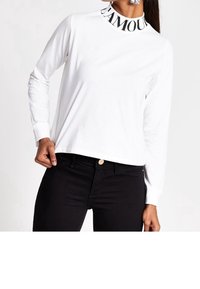 Frau in einem weißen Langarmshirt mit dem Schriftzug "GLAMOUR" am Kragen und schwarzen Hosen, posiert mit einer Hand in der Tasche.