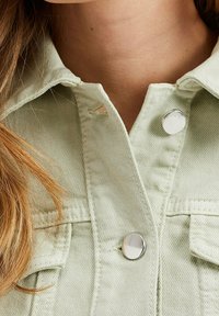 Vaaleanvihreä farkkupaita, jossa on kauluskäänne, hopeiset nepparit ja näkyvät ompeluyksityiskohdat. Hiukset peittävät osittain kaulusta.