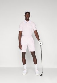Ljusrosa polo med krage och blå detaljer, tillsammans med ljusrosa shorts. Vita golfskor, vita strumpor med svarta detaljer och en keps.