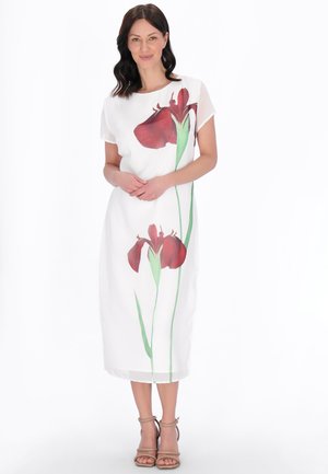 Kobieta w białej sukience midi z dużymi czerwonymi kwiatowymi nadrukami, stojąca ze złączonymi rękami, ubrana w beżowe szpilki na kostkę na białym tle.