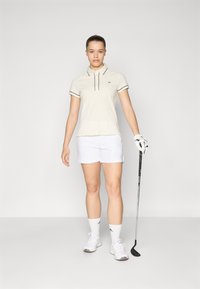 Beige golf polo med marinblå randdetaljer, vita shorts, vita strumpor och sportskor, håller en golfklubba. Ren, atletisk design.