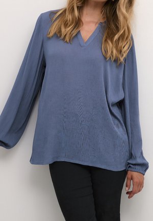 Femme portant un chemisier bleu ample à manches longues et col en V, avec un pantalon noir, debout devant un fond clair uni.