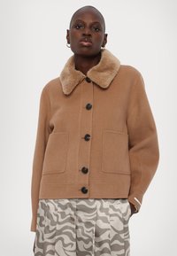 Veste en mélange de laine marron avec un col en fausse fourrure, dotée de poches avant et de boutons noirs ; associée à un pantalon plissé à motifs.