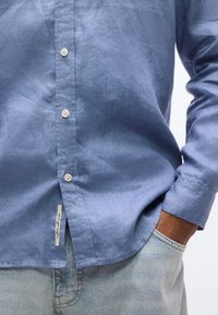 Άνδρας που φοράει γαλάζιο πουκάμισο μακρυμάνικο με κουμπιά και ετικέτα κοντά στο  κάτω μέρος, με το χέρι στην τσέπη ενός ανοιχτόχρωμου τζιν.