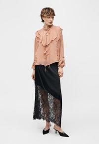 Sieviete, kas valkā persiku krāsas volānu blūzi un melnu asimetrisku svārku ar caurspīdīgu mežģīņu paneli, kopā ar melniem smailiem zemu papēžu apaviem, stāv.